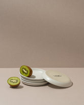 A white stoneware flat side plate with a matte finish, displayed with a halved kiwi on top, against a neutral background.